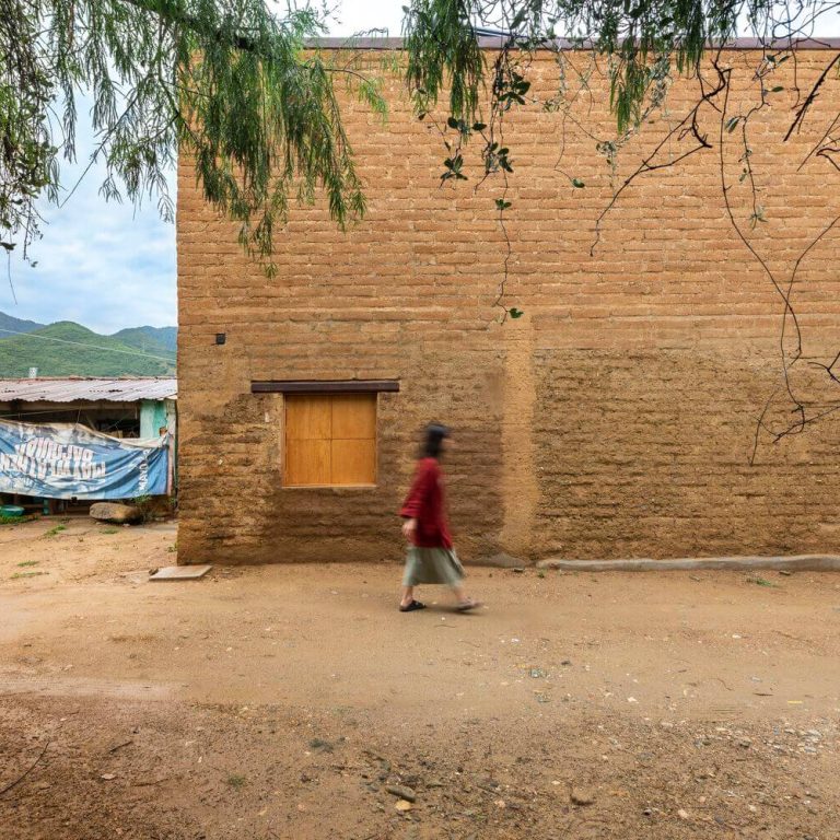 Fachada de casa de adobe con ventana de madera, una persona caminando. Casa taller Plúmula en Oaxaca por Espacio 18 Arquitectura